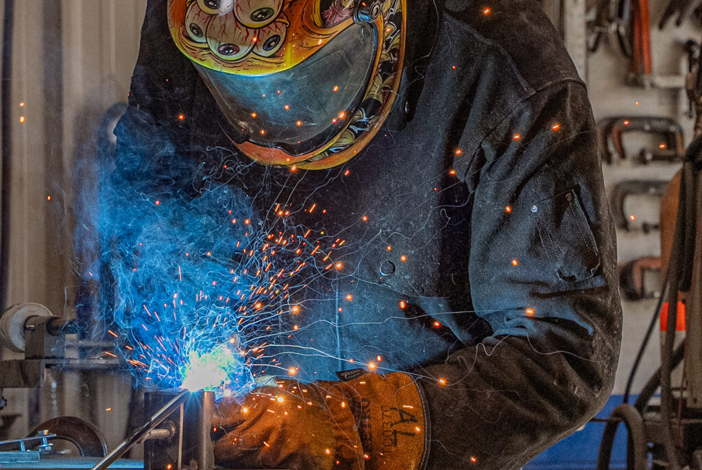 Welder wearing protective gear sparks metal with bright blue flame in a workshop. Industrial photography capturing custom welding services in Tipp City, Ohio.