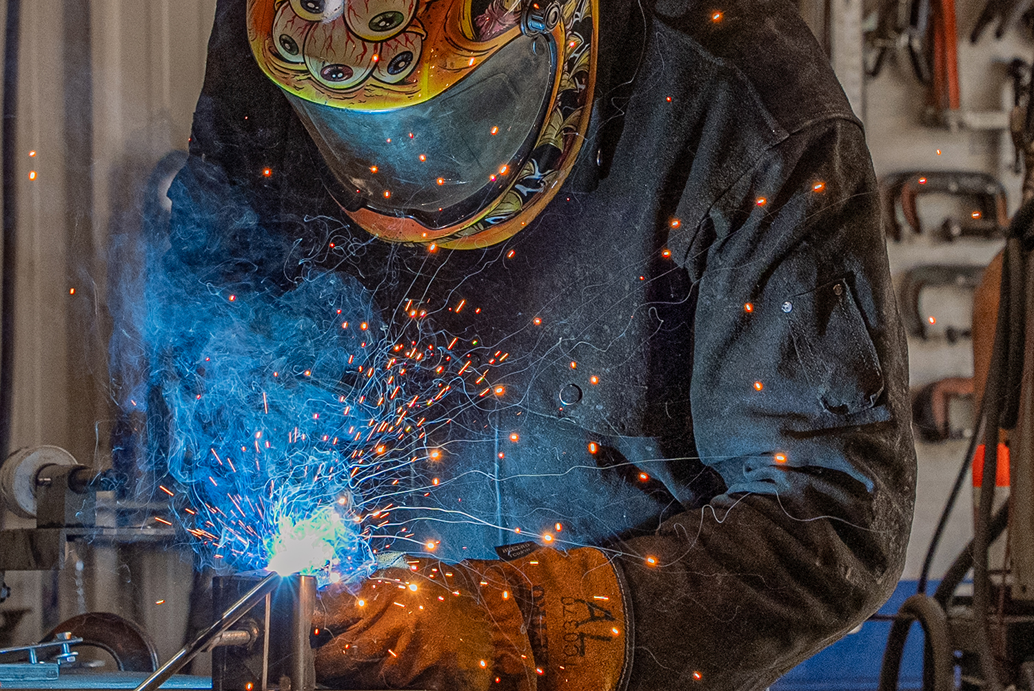 Welder wearing protective gear sparks metal with bright blue flame in a workshop. Industrial photography capturing custom welding services in Tipp City, Ohio.